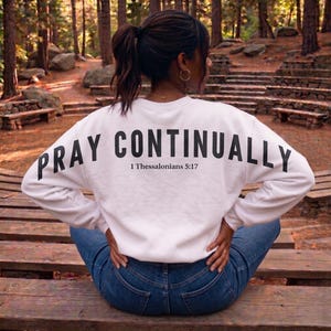 May include: White sweatshirt with the words "PRAY CONTINUALLY" printed in black across the back, along with the text "1 Thessalonians 5:17". The sweatshirt is paired with blue jeans. The image is taken outdoors.