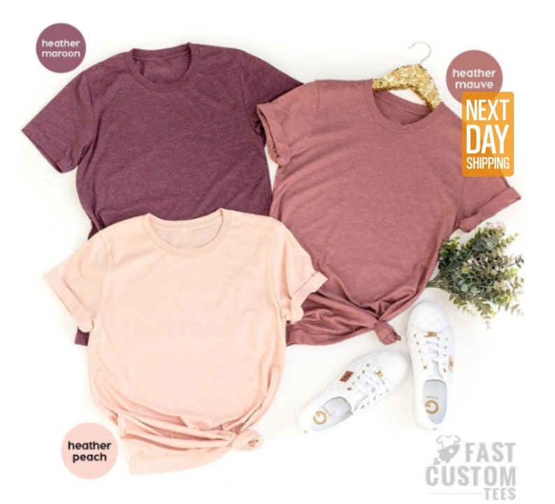 May include: Three plain t-shirts in different colors: heather maroon, heather mauve, and heather peach. The shirts are laid out on a white surface.  The text "NEXT DAY SHIPPING" is visible in the upper right corner of the image.