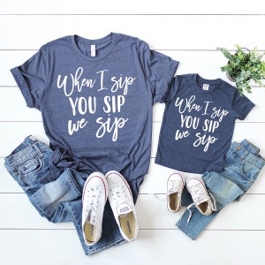 以下が含まれることがあります： 「When I sip YOU SIP we sip」と白文字で書かれた、ネイビーブルーのTシャツが2枚セットになっています。Tシャツは白い背景にジーンズと白いスニーカーと一緒に置かれています。