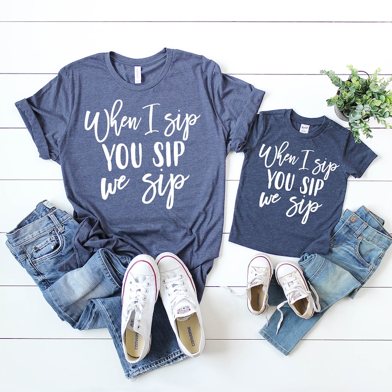 以下が含まれることがあります： 「When I sip YOU SIP we sip」と白文字で書かれた、ネイビーブルーのTシャツが2枚セットになっています。Tシャツは白い背景にジーンズと白いスニーカーと一緒に置かれています。