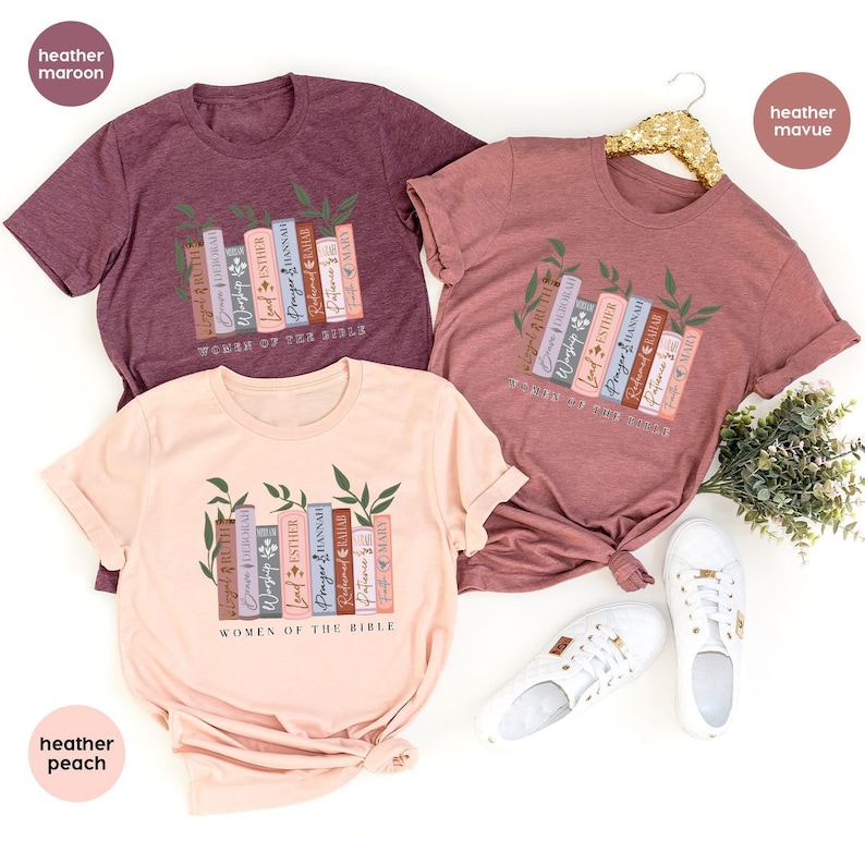 May include: Three t-shirts with a design of stacked books with names of women from the Bible. The shirts are in heather maroon, heather mauve, and heather peach.