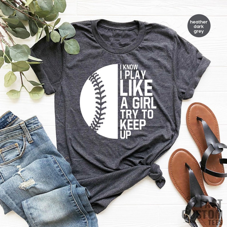 Op de afbeelding: Een donkergrijs heather t-shirt met een witte baseball-afbeelding en de tekst "I know I play like a girl try to keep up".