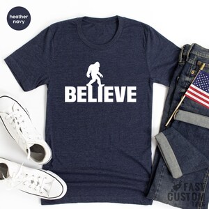 May include: A heather navy blue t-shirt with a white silhouette of Bigfoot walking and the word "BELIEVE" printed below it.