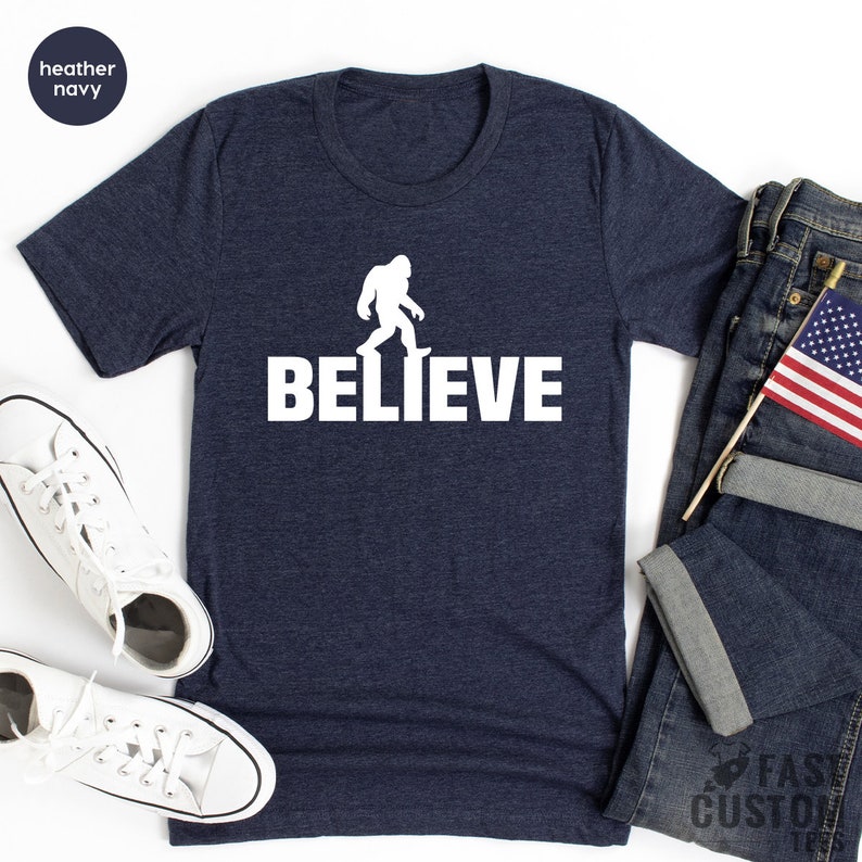 May include: A heather navy blue t-shirt with a white silhouette of Bigfoot walking and the word "BELIEVE" printed below it.