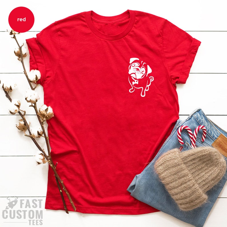 May include: Red t-shirt with a white pug graphic on the front. The shirt is laid flat on a white surface with a cotton branch, blue jeans, a brown hat, and candy canes.