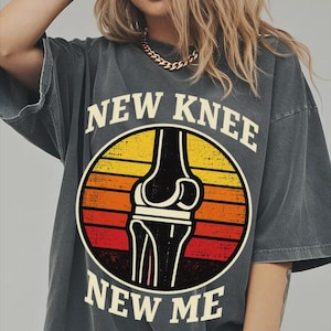 May include: A charcoal gray short-sleeve t-shirt featuring a circular graphic with a sunset-inspired background of yellow, orange, and red stripes. The graphic depicts a black knee joint illustration with the text NEW KNEE NEW ME in cream.