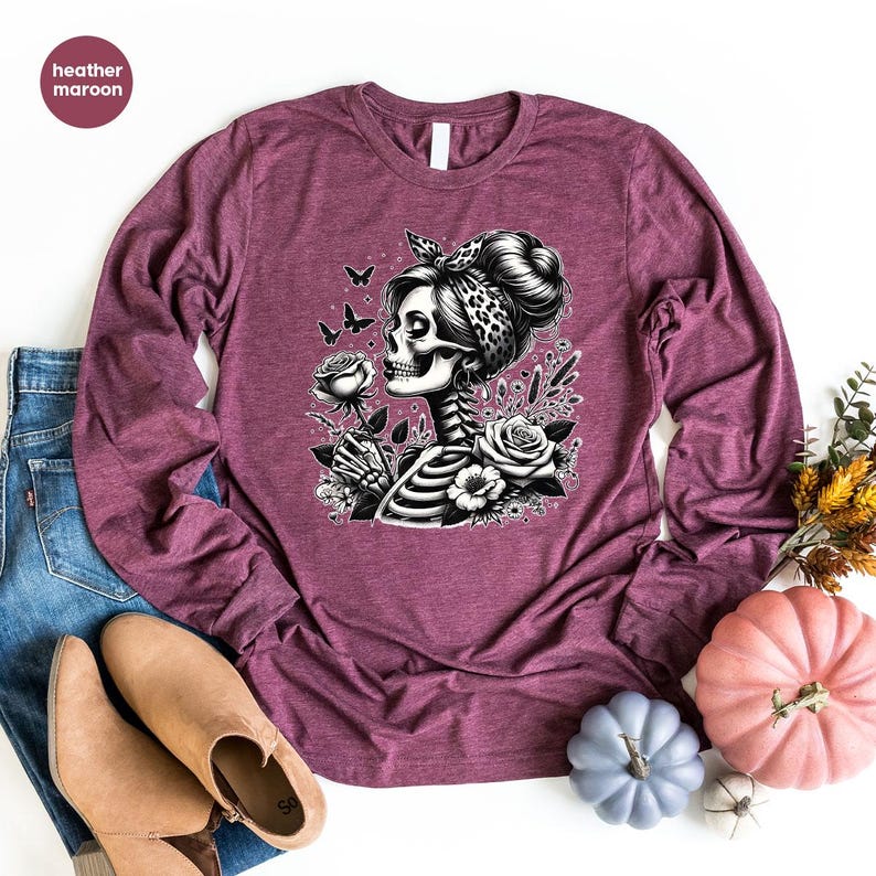 Puede incluir: Una camiseta de manga larga de color granate jaspeado con una ilustraci&oacute;n en blanco y negro de una mujer esqueleto con una rosa y mariposas. La mujer lleva una bandana de estampado de leopardo.