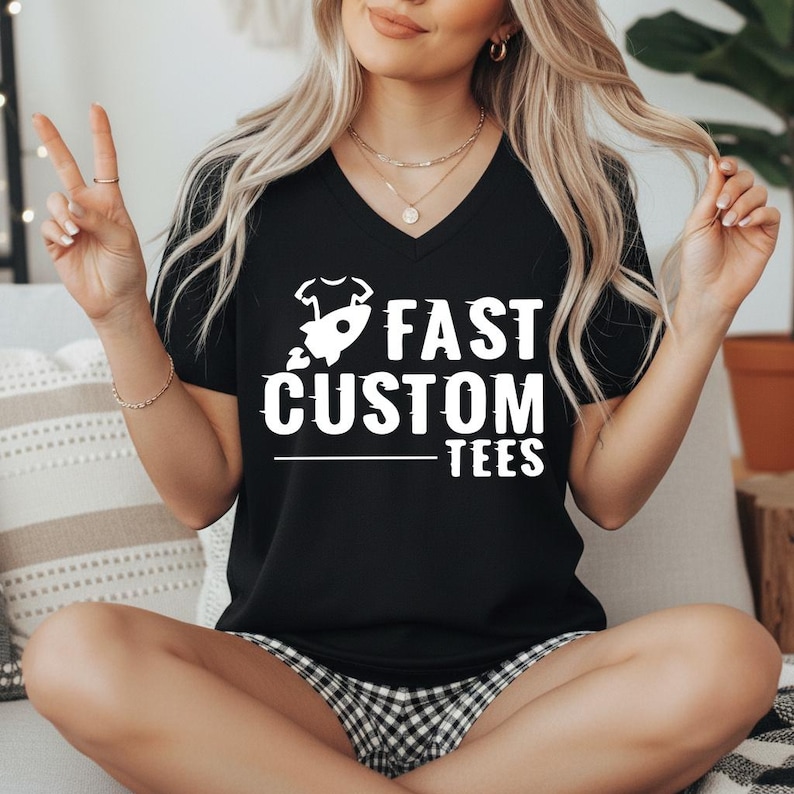 May include: Black V-neck t-shirt with the text "FAST CUSTOM TEES" in white. The design includes a rocket ship and a baby onesie. The shirt is a casual wear item.