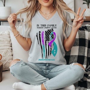 以下が含まれることがあります： ライトグレーのVネックTシャツ。ダメージ加工されたアメリカ国旗と、紫とティールブルーのリボンが描かれています。黒とティールブルーの文字で「IN THIS FAMILY NOBODY FIGHTS ALONE」「SUICIDE PREVENTION」「AWARENESS」とプリントされています。
