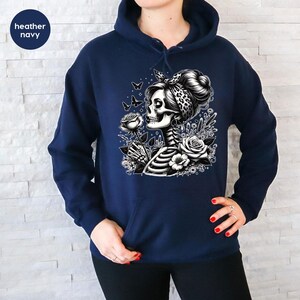 Puede incluir: Una sudadera con capucha azul marino con un gr&aacute;fico blanco de una mujer esqueleto sosteniendo una rosa, rodeada de mariposas y flores.