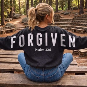May include: A dark gray sweatshirt with the word "FORGIVEN" in large white letters across the back, with "Psalm 32:1" below. The sweatshirt is worn with blue jeans. The setting is outdoors, with trees and wooden benches.