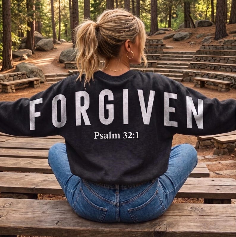 May include: A dark gray sweatshirt with the word "FORGIVEN" in large white letters across the back, with "Psalm 32:1" below. The sweatshirt is worn with blue jeans. The setting is outdoors, with trees and wooden benches.