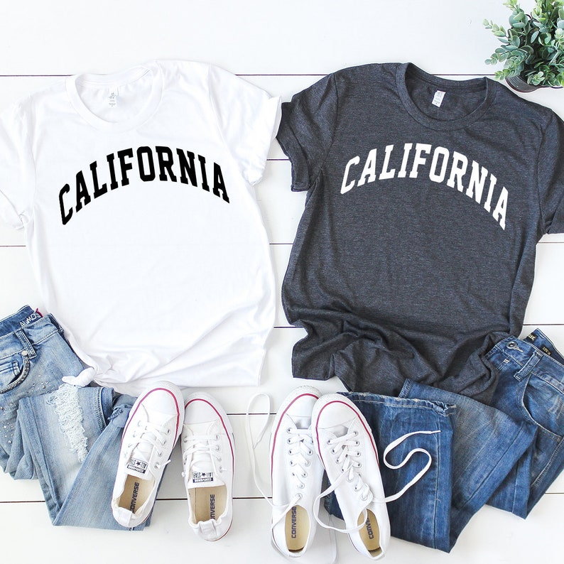 May include: Two t-shirts with the word "CALIFORNIA" printed in a bold, arched font. One shirt is white with black text, and the other is dark gray with white text. Both shirts are laid out on a white wooden surface with a pair of white sneakers and blue jeans.