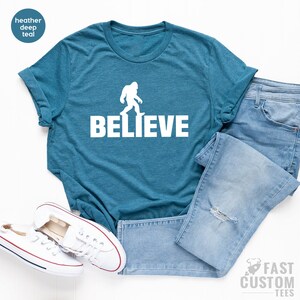 May include: A teal blue t-shirt with a white silhouette of Bigfoot walking and the word "BELIEVE" printed below it.