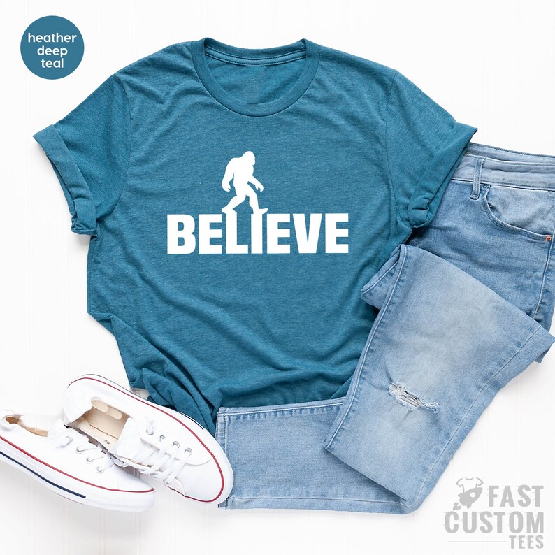 May include: A teal blue t-shirt with a white silhouette of Bigfoot walking and the word "BELIEVE" printed below it.