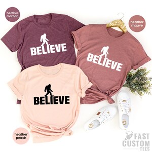 May include: Three t-shirts with a Bigfoot graphic and the word "BELIEVE" printed on them. The shirts are in heather maroon, heather mauve, and heather peach.