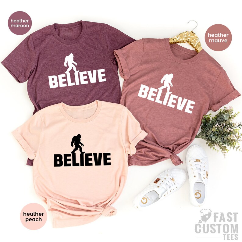 May include: Three t-shirts with a Bigfoot graphic and the word "BELIEVE" printed on them. The shirts are in heather maroon, heather mauve, and heather peach.