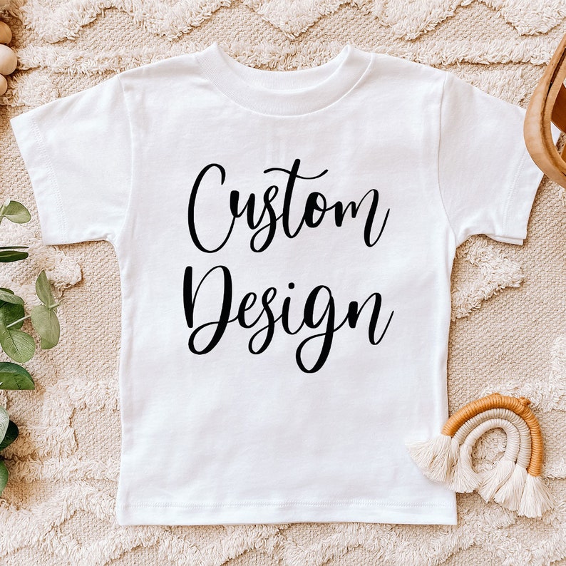 May include: White t-shirt with the text "Custom Design" printed in black cursive font.