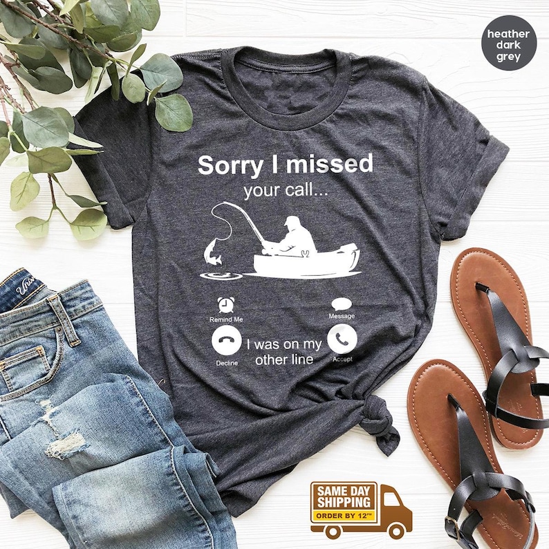 May include: A heather dark grey t-shirt with a white graphic of a phone screen. The phone screen shows a missed call notification with the text 'Sorry I missed your call...' and a cartoon image of a person fishing in a boat. The phone screen also shows a button that says 'I was on my other line'. The t-shirt is displayed on a white surface with a pair of brown sandals and a pair of blue jeans.