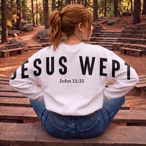 May include: A white sweatshirt with the words "JESUS WEPT" printed in large black letters across the back, with "John 11:35" below. The person is wearing blue jeans and sitting on a wooden bench.