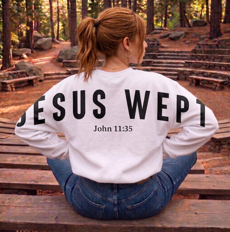 May include: A white sweatshirt with the words "JESUS WEPT" printed in large black letters across the back, with "John 11:35" below. The person is wearing blue jeans and sitting on a wooden bench.
