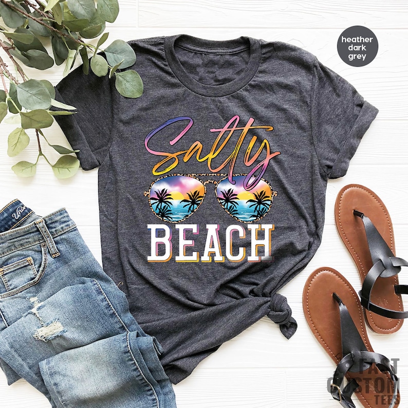 May include: A heather dark grey t-shirt with the text "Salty Beach" in rainbow colors. The text is surrounded by a pair of sunglasses with a sunset and palm trees reflected in the lenses.
