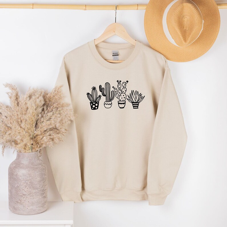 May include: A beige sweatshirt featuring a black graphic of four potted cacti. The cacti are in different shapes and sizes, with detailed pots. The sweatshirt is hanging on a wooden hanger, with a straw hat in the background.