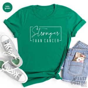 May include: A green t-shirt with the text "Stronger Than Cancer" in white script within a white rectangle. The image includes grey trainers and denim shorts.