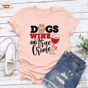 May include: Heather peach-colored t-shirt with the words "Dogs, Wine, and True Crime" in a graphic design. The design includes a paw print, a wine glass, and a splash of red. The shirt is paired with denim shorts and white sneakers.