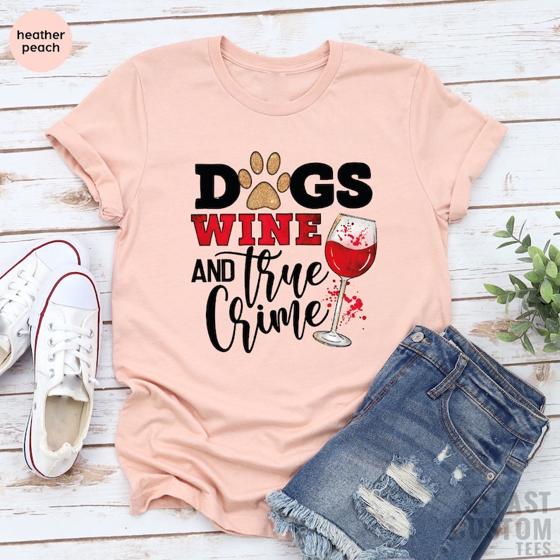 May include: Heather peach-colored t-shirt with the words "Dogs, Wine, and True Crime" in a graphic design. The design includes a paw print, a wine glass, and a splash of red. The shirt is paired with denim shorts and white sneakers.