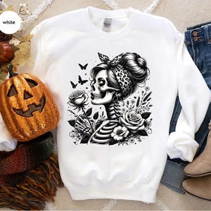 Puede incluir: Sudadera blanca con una ilustraci&oacute;n en blanco y negro de una mujer esqueleto sosteniendo una flor, con una bandana en la cabeza y rodeada de flores.