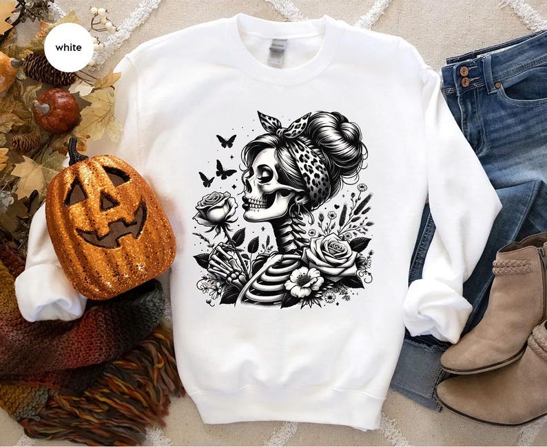 Puede incluir: Sudadera blanca con una ilustraci&oacute;n en blanco y negro de una mujer esqueleto sosteniendo una flor, con una bandana en la cabeza y rodeada de flores.