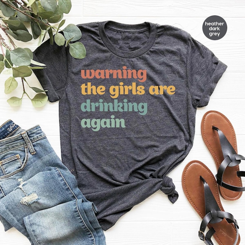 Pode incluir: Uma camiseta cinza escura mesclada com o texto "warning the girls are drinking again" em letras retr&ocirc; coloridas.