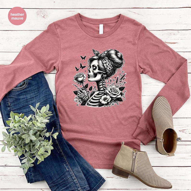 Puede incluir: Una camiseta de manga larga de color malva jaspeado con un gr&aacute;fico de una mujer esqueleto con una rosa en la mano y mariposas a su alrededor. El gr&aacute;fico es en blanco y negro y est&aacute; rodeado de flores.