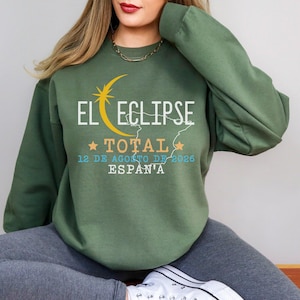 Peut inclure: Sweat-shirt vert avec l'inscription "EL ECLIPSE TOTAL 12 DE AGOSTO DE 2026 ESPAÑA" en blanc, jaune et orange. Le design comprend un croissant de lune et une étoile. Le sweat-shirt est de style col rond.