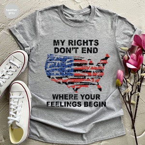 May include: Grey t-shirt featuring the text "MY RIGHTS DON'T END WHERE YOUR FEELINGS BEGIN" and a distressed American flag design. White canvas trainers are visible on the left. The shirt is folded and displayed with a floral arrangement.