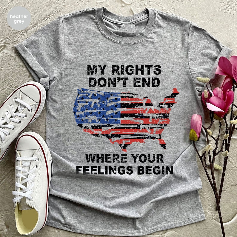 May include: Grey t-shirt featuring the text "MY RIGHTS DON'T END WHERE YOUR FEELINGS BEGIN" and a distressed American flag design. White canvas trainers are visible on the left. The shirt is folded and displayed with a floral arrangement.