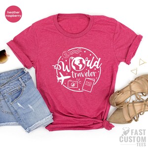 May include: A heather raspberry t-shirt with a white graphic design of a world with the text "World Traveler" and a plane, camera, and passport.