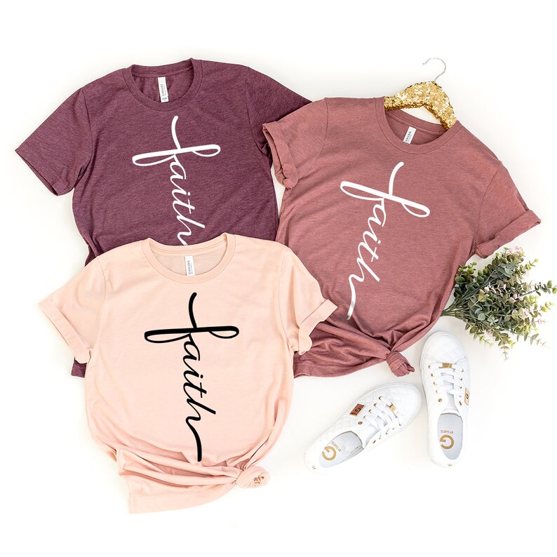 May include: Three t-shirts with the word "faith" written in a cursive font on a cross design. The shirts are in different shades of pink and burgundy.