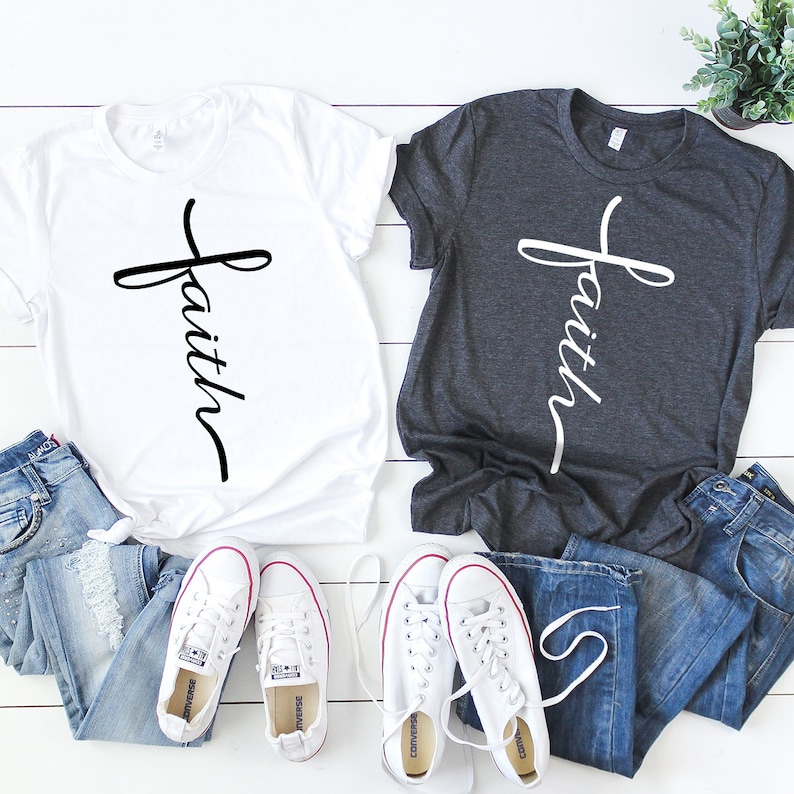 May include: Two t-shirts with the word "faith" written vertically on a cross design. One shirt is white and the other is dark gray. Both shirts are laid out on a white wooden surface with a pair of white sneakers on each side.