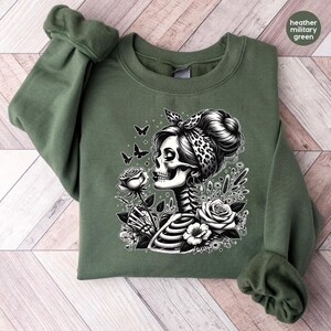 Puede incluir: Una sudadera verde con un estampado gr&aacute;fico en blanco y negro de una mujer esqueleto sosteniendo una rosa y rodeada de mariposas. Lleva una bandana de estampado de leopardo.