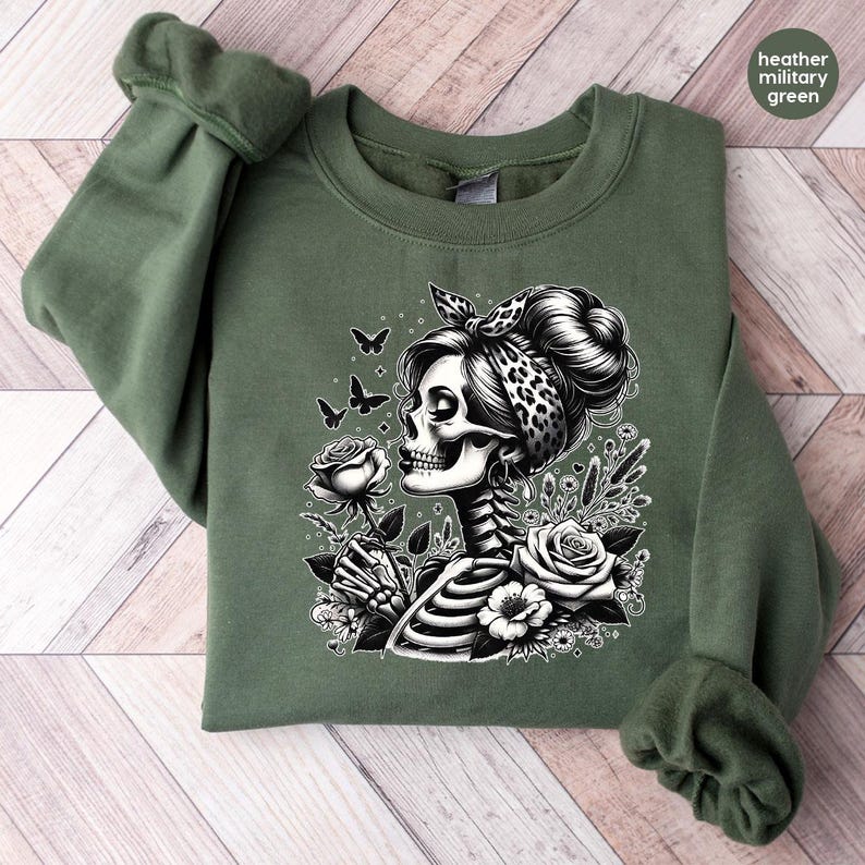 Puede incluir: Una sudadera verde con un estampado gr&aacute;fico en blanco y negro de una mujer esqueleto sosteniendo una rosa y rodeada de mariposas. Lleva una bandana de estampado de leopardo.