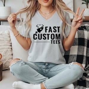 May include: A light gray V-neck t-shirt with the text "FAST CUSTOM TEES" in black. The graphic includes a rocket ship and a baby onesie. The model is wearing light blue jeans.