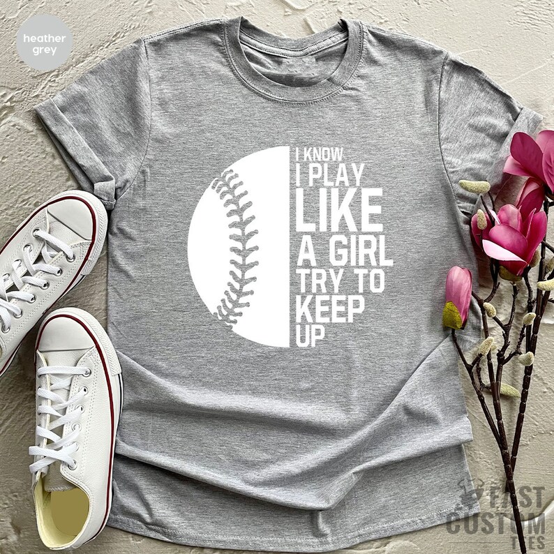 Op de afbeelding: Heather grijs T-shirt met een witte baseball graphic en de tekst "I know I play like a girl try to keep up".
