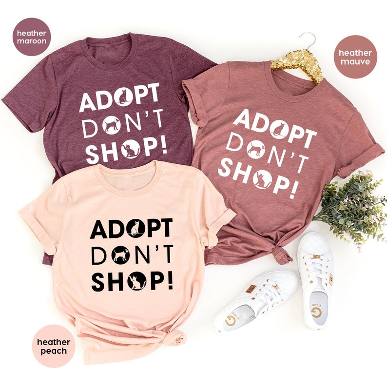 May include: Three t-shirts in different colors with the text "Adopt Don't Shop!" printed on them. The shirts are heather maroon, heather peach, and heather mauve.