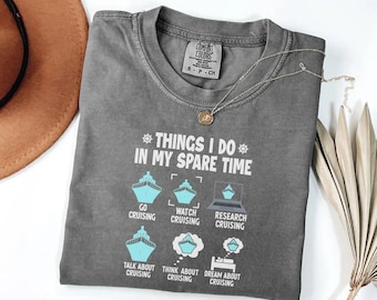 May include: A gray Comfort Colors t-shirt with the text "THINGS I DO IN MY SPARE TIME" and cruise ship graphics. The shirt also says "GO CRUISING", "WATCH CRUISING", "RESEARCH CRUISING", "TALK ABOUT CRUISING", "THINK ABOUT CRUISING", and "DREAM ABOUT CRUISING". A gold necklace is on top of the shirt.