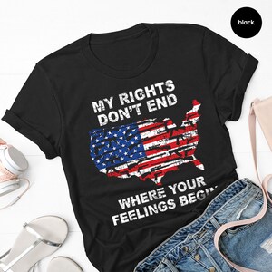 May include: Black t-shirt featuring the text "MY RIGHTS DON'T END WHERE YOUR FEELINGS BEGIN" above a distressed American flag design shaped like the USA. The shirt is folded and displayed on a white surface.