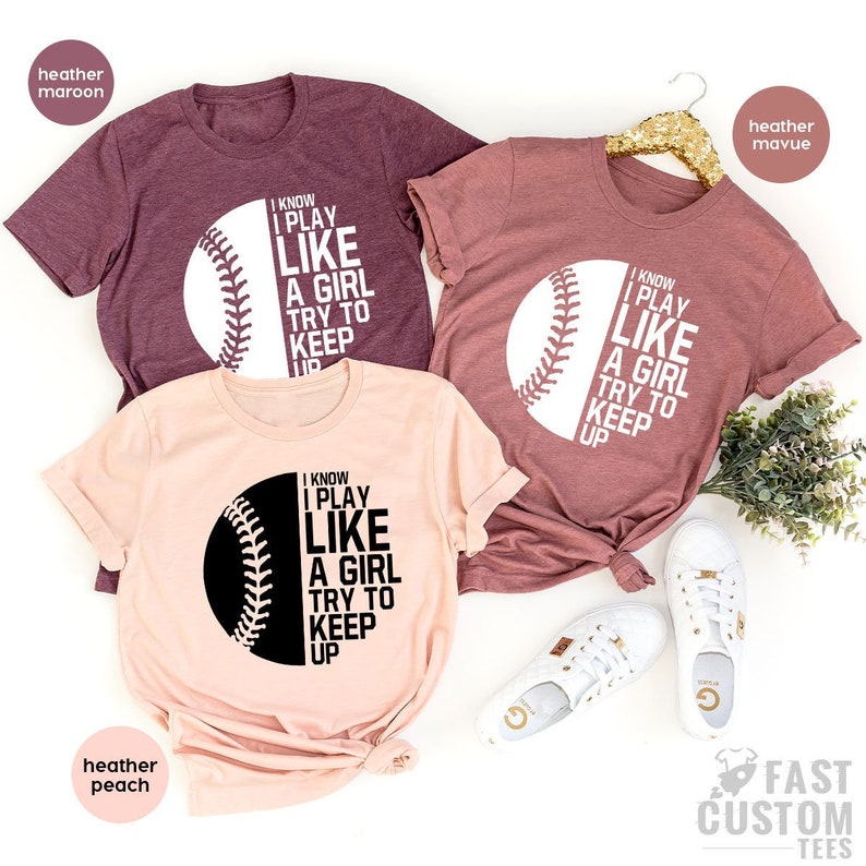 Op de afbeelding: Drie T-shirts met een baseball-afbeelding en de tekst "Ik weet dat ik speel als een meisje, probeer bij te blijven". De shirts zijn in heather maroon, heather peach en heather mauve.