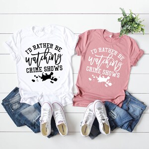 Pode incluir: Uma t-shirt branca com texto preto e uma t-shirt malva com texto branco, ambas com a frase I'D RATHER BE watching CRIME SHOWS e um gr&aacute;fico de salpicos de tinta. Cada t-shirt &eacute; combinada com cal&ccedil;as de ganga azuis e t&eacute;nis brancos.