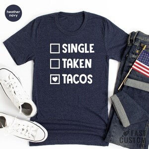 May include: A heather navy blue t-shirt with a white graphic that reads "Single, Taken, Tacos" with a heart replacing the checkmark for the "Tacos" option.